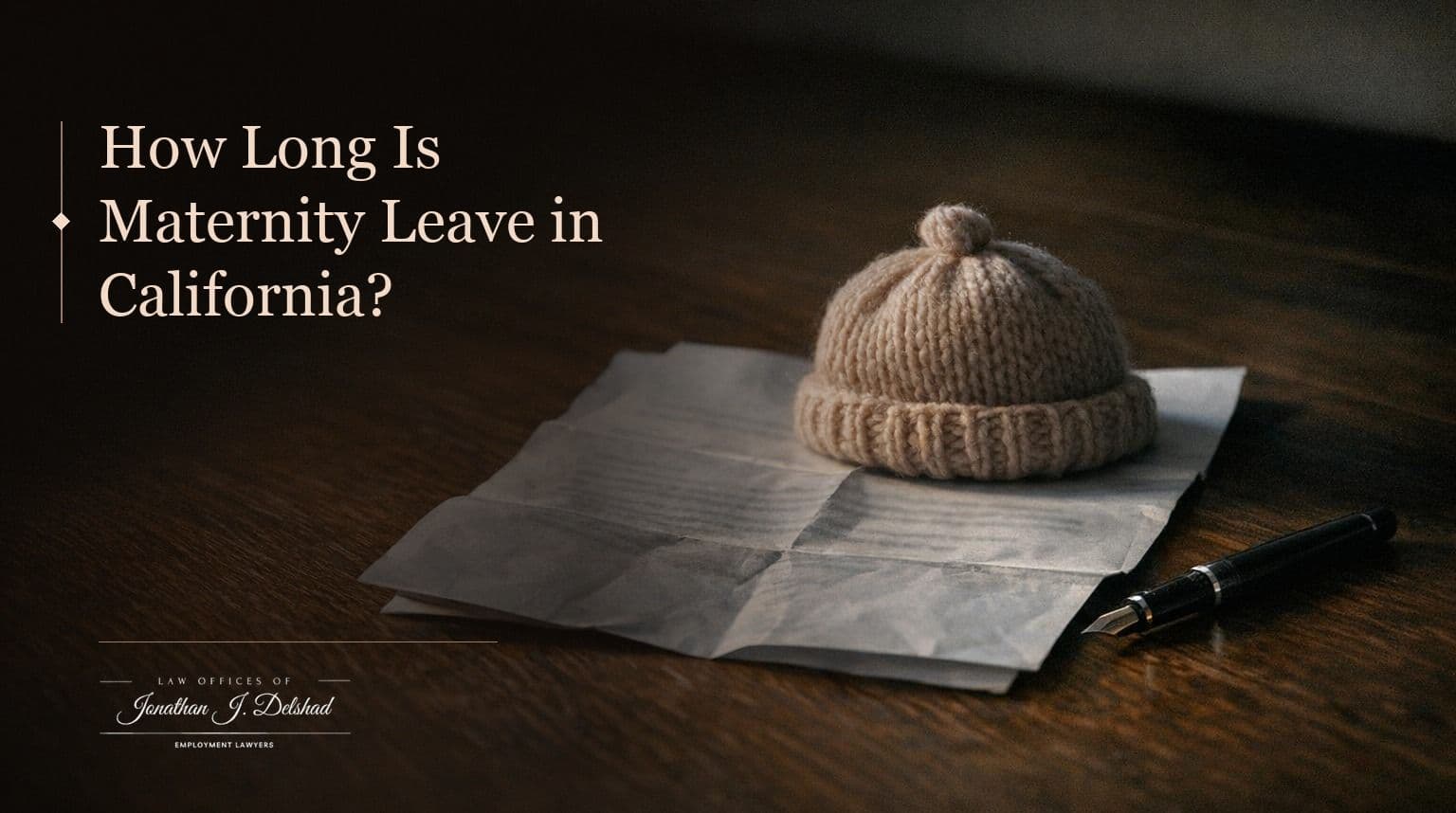 A small knitted infant hat resting on a folded paper form on a dark walnut table beside a fountain pen, headed 'How Long Is Maternity Leave in California?'